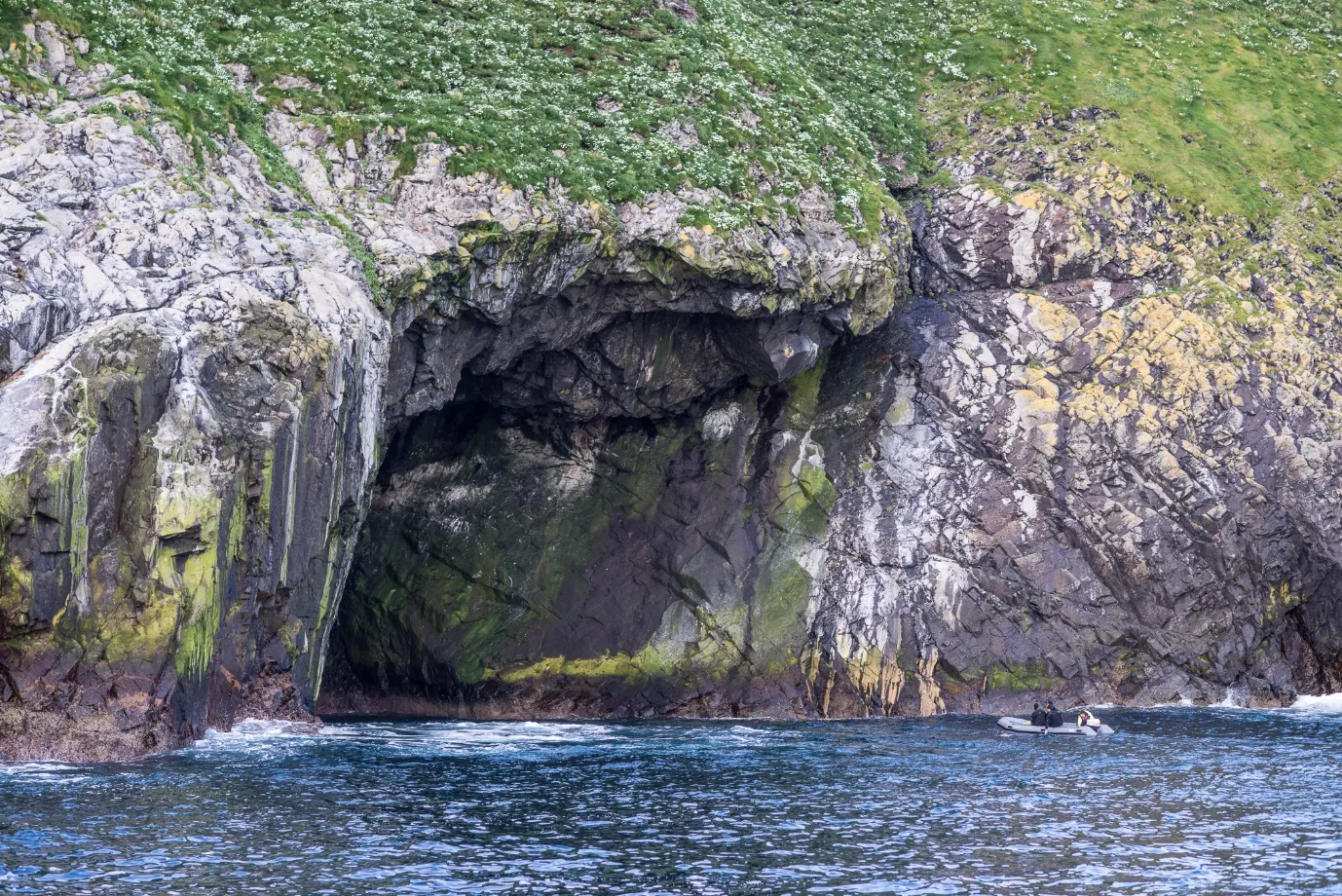 A large sea cave entrance at the base of a grassy cliff